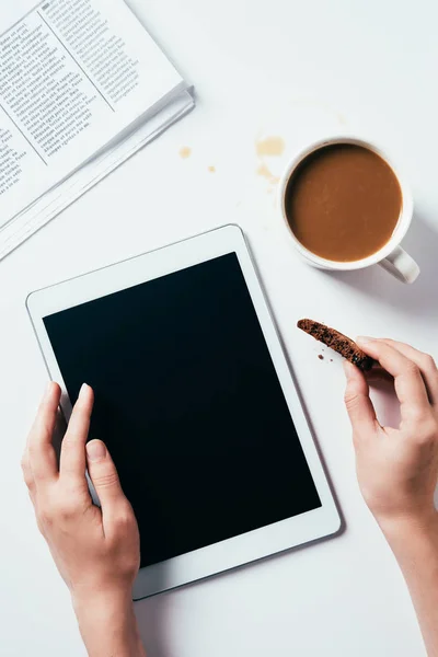 Tiro recortado de la mujer que usa la tableta mientras come galletas de chocolate con café en la superficie blanca - foto de stock