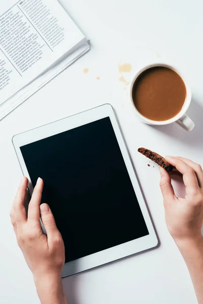 Abgeschnittene Aufnahme einer Frau mit Tablette beim Essen von Schokoladenkeksen mit Kaffee auf weißer Oberfläche — Stockfoto