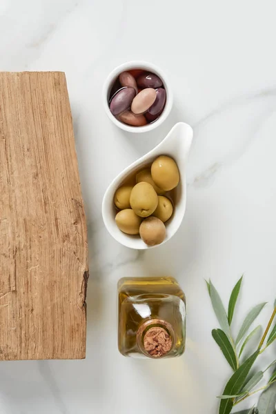 Top view of bottle of olive oil, log and olives in bowls on marble table — Stock Photo