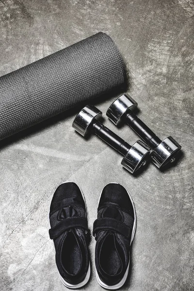 High angle view of view of sporting shoes with yoga mat and dumbbells lying on concrete surface — Stock Photo