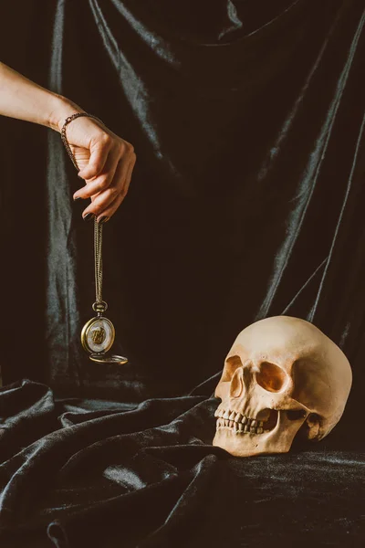 Cropped view of woman holding vintage clock on the chain near skull on black cloth — Stock Photo