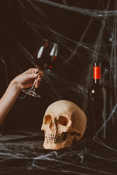 Cropped view of woman holding glass of red wine near skull on black cloth with spider web — Stock Photo