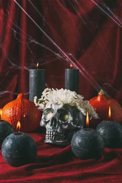 Cráneo de halloween plateado con flores, velas negras y calabazas en tela roja con tela de araña - foto de stock