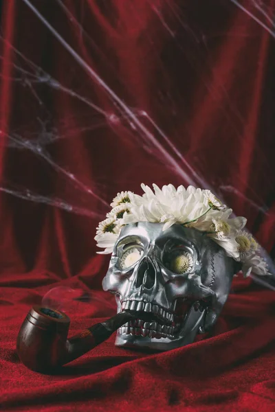 Crâne d'argent avec des fleurs et pipe à fumer sur tissu rouge avec toile d'araignée — Photo de stock
