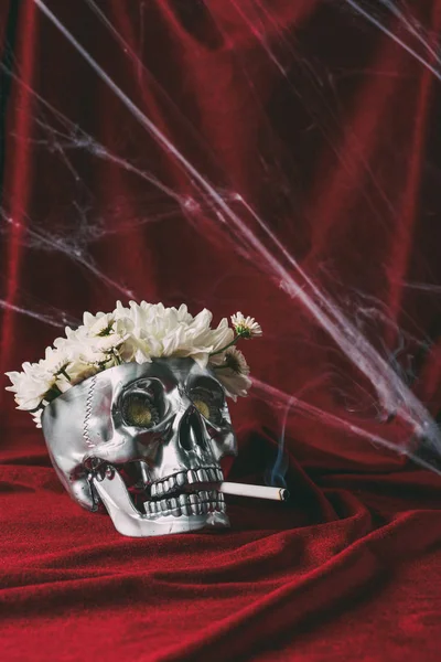 Silver skull with flowers smoking cigarette on red cloth with spider web — Stock Photo