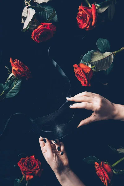 Cropped shot of woman holding black mask and beautiful red roses on black — Stock Photo