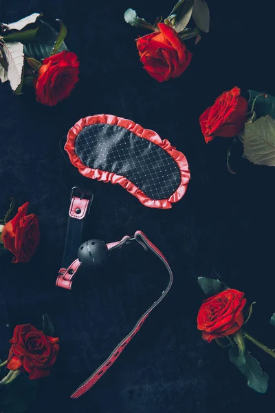 Vue du haut du masque pour les yeux, bâillon et roses rouges sur noir — Photo de stock