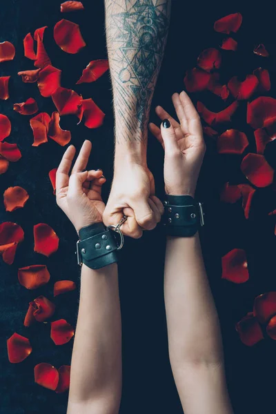 Cropped shot of man holding handcuffs on female hands above black fabric with red rose petals — Stock Photo