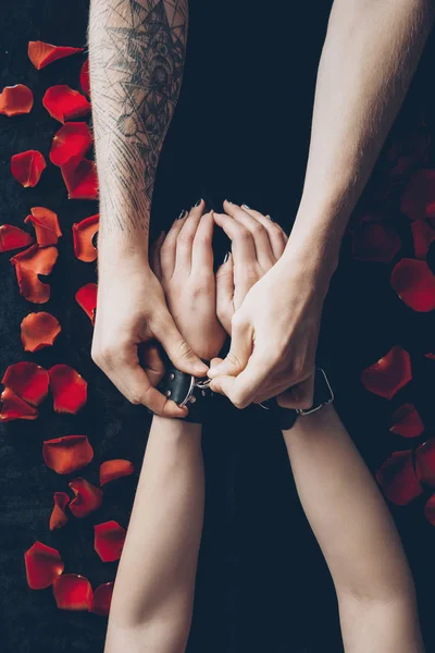 Cropped shot of couple with leather handcuffs above black fabric with red rose petals — Stock Photo