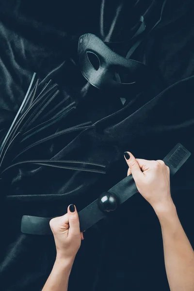 Plan recadré de mains féminines avec bâillon, fouet en cuir et masque sur tissu noir — Photo de stock