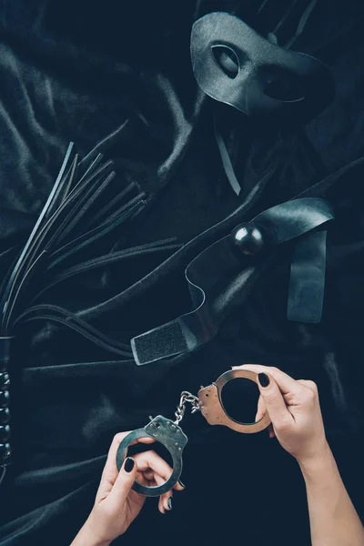 Cliché recadré de femme tenant menottes, fouet en cuir, bâillon et masque sur tissu noir — Photo de stock