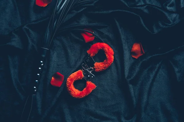 Draufsicht auf rote flauschige Handschellen, Lederpeitsche und Rosenblätter auf schwarzem Stoff — Stockfoto