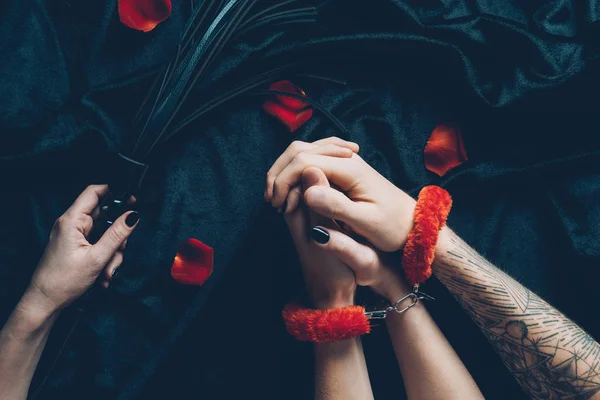 Cropped shot of couple in red fluffy handcuffs holding black leather whip — Stock Photo