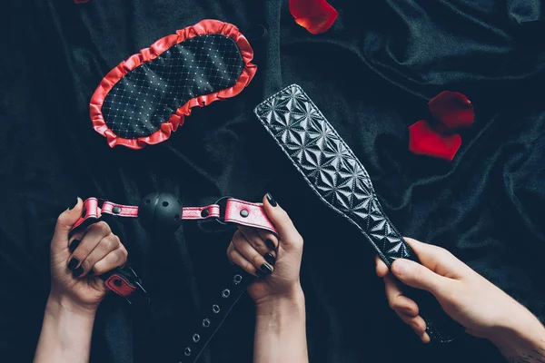 Cropped shot of couple holding black leather spanking paddle and gag — Stock Photo