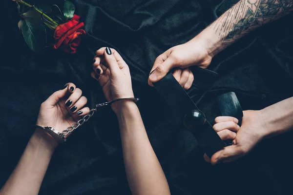 Tiro recortado de pareja con esposas y mordaza sobre tela negra con flor de rosa - foto de stock