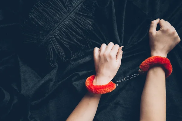 Cropped shot of woman in red fluffy handcuffs holding black fabric with feather — Stock Photo