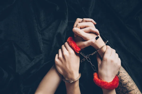 Cropped shot of couple in metal and red fluffy handcuffs on black fabric — Stock Photo