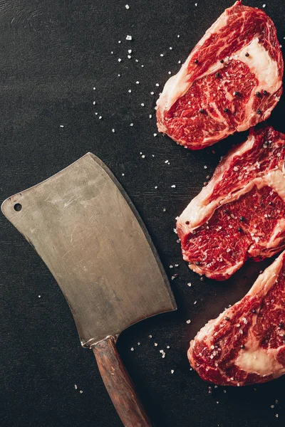 Вид сверху на стейки из сырого мяса и тесак на поверхности на кухне — стоковое фото