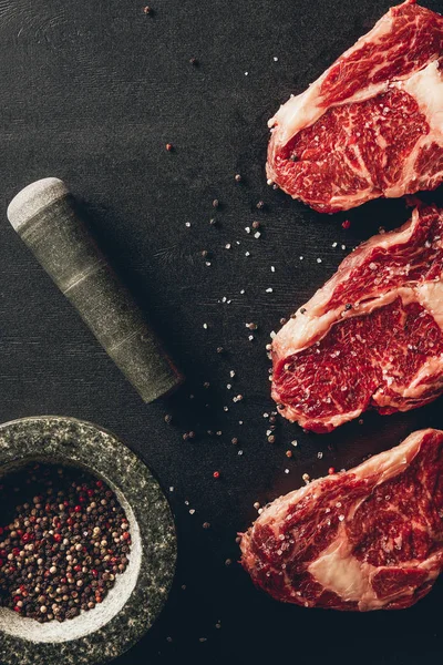 Top view of raw meat steaks, mortar, pestle and pepper on table in kitchen — Stock Photo