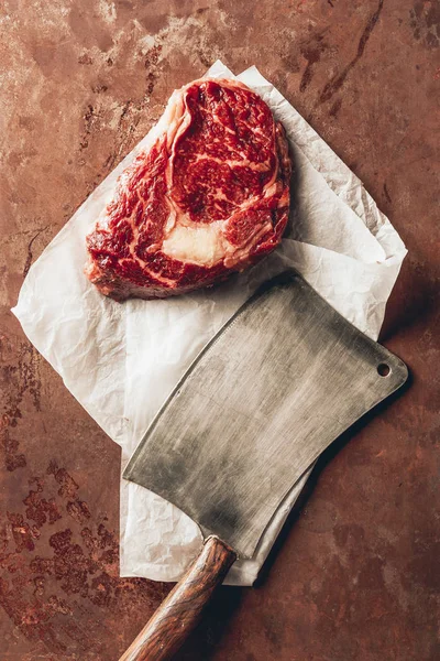 Top view of raw meat steak, baking paper and cleaver on tabletop in kitchen — Stock Photo