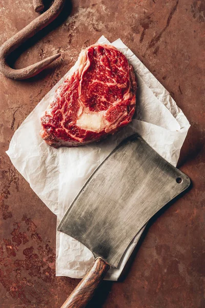 Top view of raw meat steak, metal hook and cleaver on tabletop in kitchen — Stock Photo