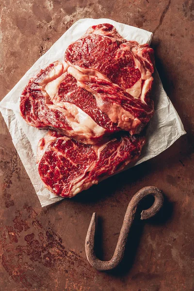 Vista superior de bifes de carne crua e gancho de metal na superfície marrom na cozinha — Fotografia de Stock