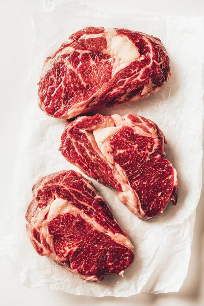 Vista superior de tres trozos de filetes de carne cruda sobre papel de hornear en la cocina - foto de stock