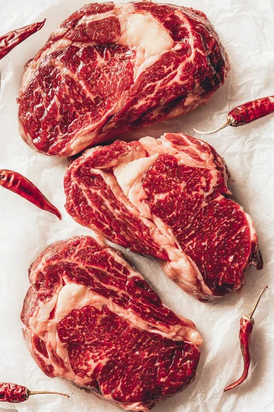 Vista dall'alto di tre pezzi di bistecche di carne cruda e peperoncino su carta da forno in cucina — Foto stock