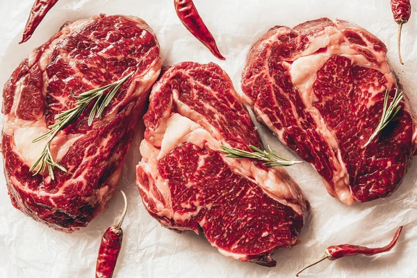 Vue de dessus de trois morceaux de viande crue steaks, piments et romarin sur papier cuisson dans la cuisine — Photo de stock