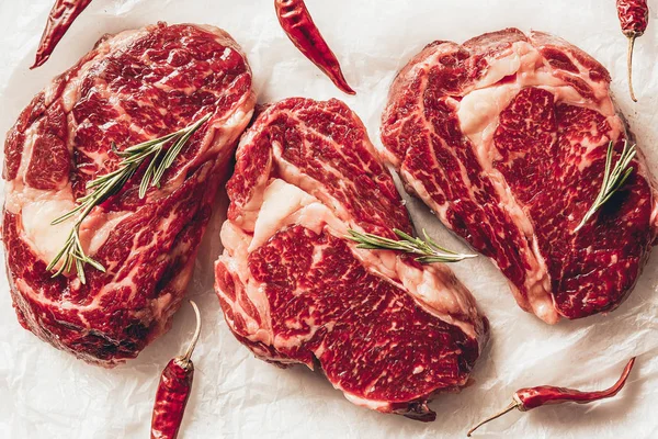 Top view of three pieces of raw meat steaks, chili peppers and rosemary on baking paper in kitchen — Stock Photo