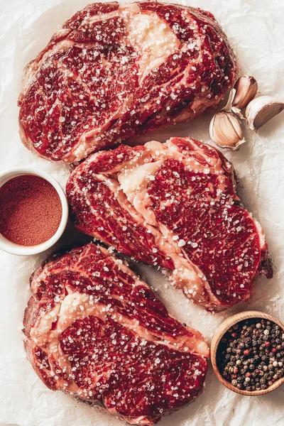 Vue de dessus de trois morceaux de steaks de viande crue avec des épices et de l'ail sur du papier cuisson dans la cuisine — Photo de stock
