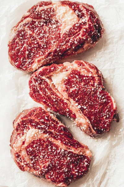 Vue de dessus de trois morceaux de steaks de viande crue avec du sel sur du papier cuisson dans la cuisine — Photo de stock