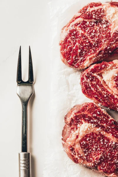 Elevated view of three pieces of raw meat steaks and meat fork on table in kitchen — Stock Photo