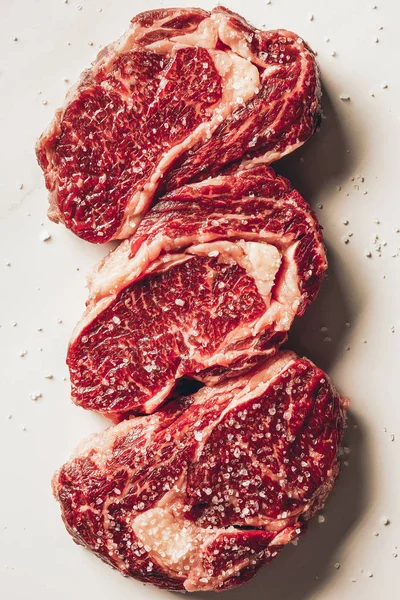 Top view of three pieces of raw meat steaks and salt on white surface — Stock Photo