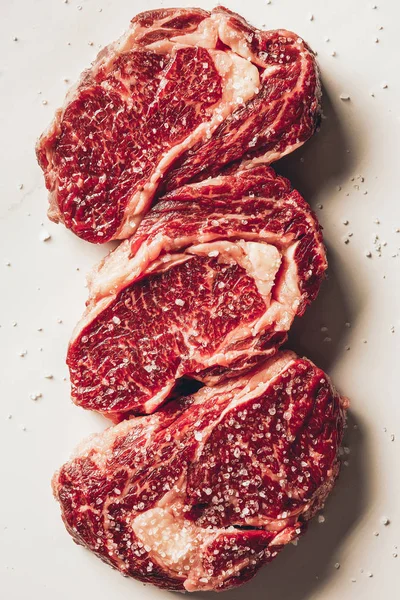 Vista superior de tres trozos de filetes de carne cruda y sal en la superficie blanca - foto de stock