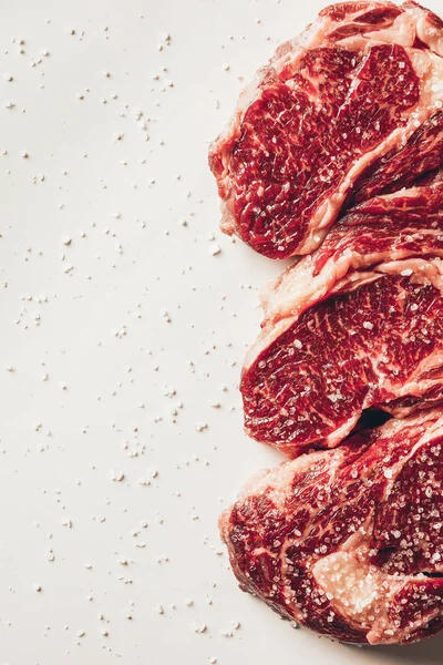 Vista elevada de tres trozos de filetes de carne cruda y sal dispersa en la superficie blanca - foto de stock