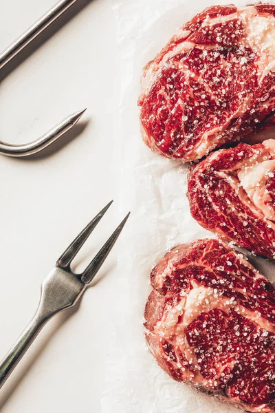 Vista dall'alto di tre pezzi di carne cruda bistecche con sale, forchetta e gancio in metallo su superficie bianca — Foto stock