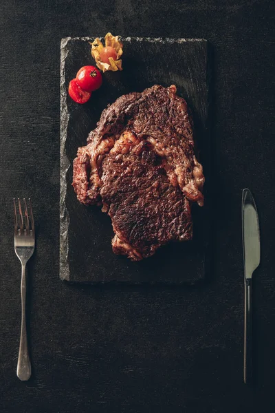 Top view of cooked steak and cherry tomato on black wooden board, knife and fork on table in kitchen — Stock Photo