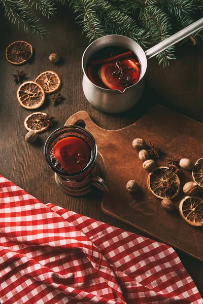 Homemade hot spiced wine in glass cup and saucepan with dried orange slices, nutmeg seeds and anise stars on wooden background — Stock Photo