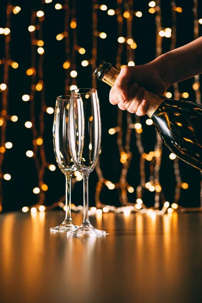 Cropped image of woman holding champagne bottle near glasses on garland light background, christmas concept — Stock Photo