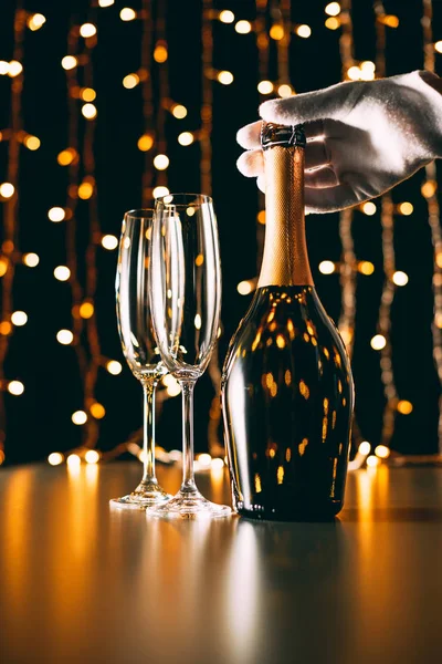 Image recadrée de serveur en gant touchant bouteille de champagne sur fond de guirlande lumière, concept de Noël — Photo de stock