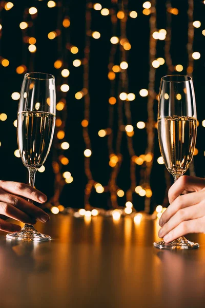 Imagen recortada de pareja sosteniendo copas de champán sobre fondo claro guirnalda, concepto de Navidad - foto de stock