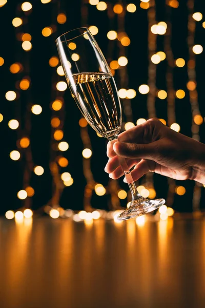 Обрезанное изображение женщины, держащей бокал шампанского на светлом фоне гирлянды, рождественская концепция — стоковое фото