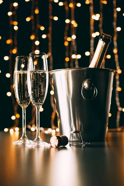 Champagne bottle in bucket and glasses on garland light background, christmas concept — Stock Photo