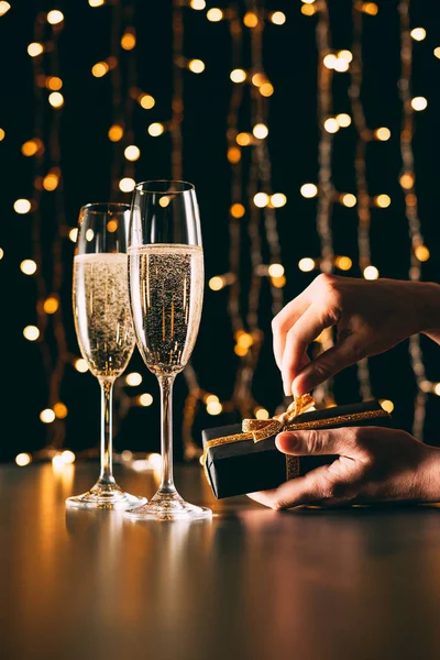 Immagine ritagliata di donna apertura presente vicino bicchieri di champagne su sfondo luce ghirlanda, concetto di Natale — Foto stock