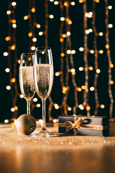 Glasses of champagne, gift and bauble on garland light background, christmas concept — Stock Photo