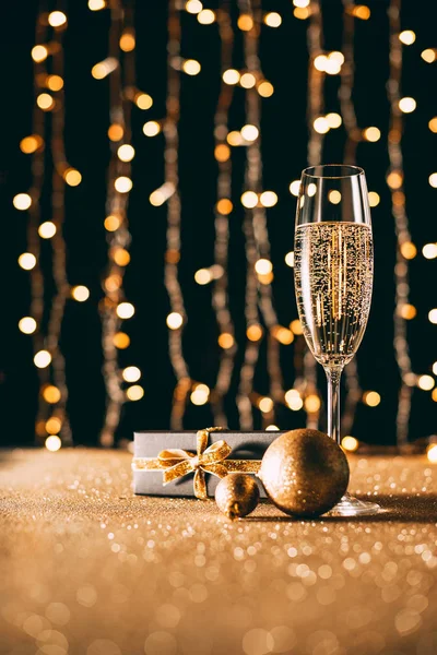 Glass of champagne, golden baubles and present on garland light background, christmas concept — Stock Photo