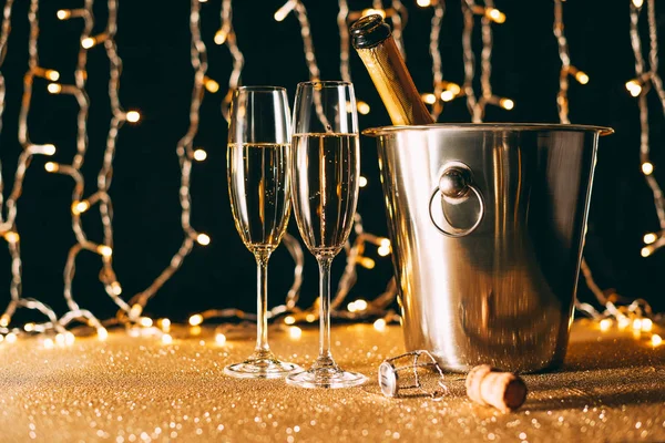 Champagne bottle in bucket and two glasses on garland light background, christmas concept — Stock Photo