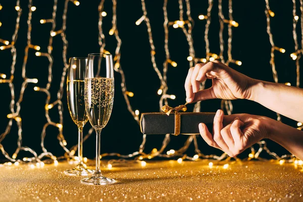 Cropped image of woman opening christmas present near champagne in glasses on garland light background — Stock Photo