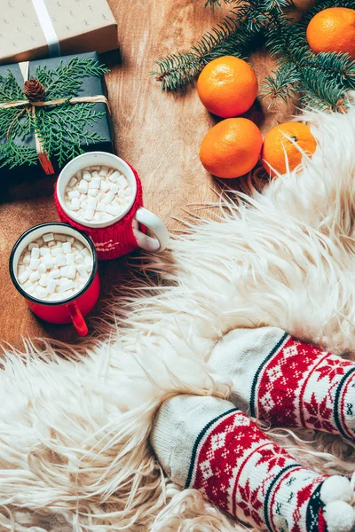 Vue partielle de la femme en chaussettes sur fond avec des tasses de boisson au cacao, mandarines, biscuits et cadeaux de Noël — Photo de stock